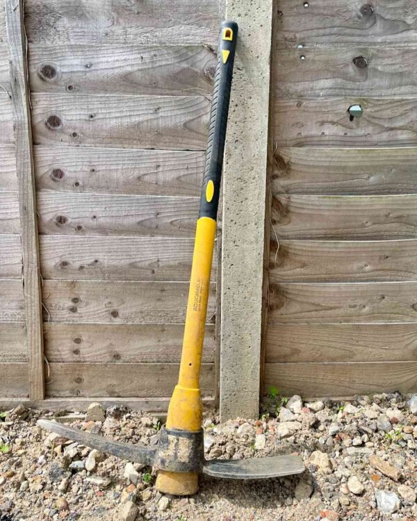 Black and yellow Roughneck 4.5lb pick mattock tool resting on a garden excavation site.