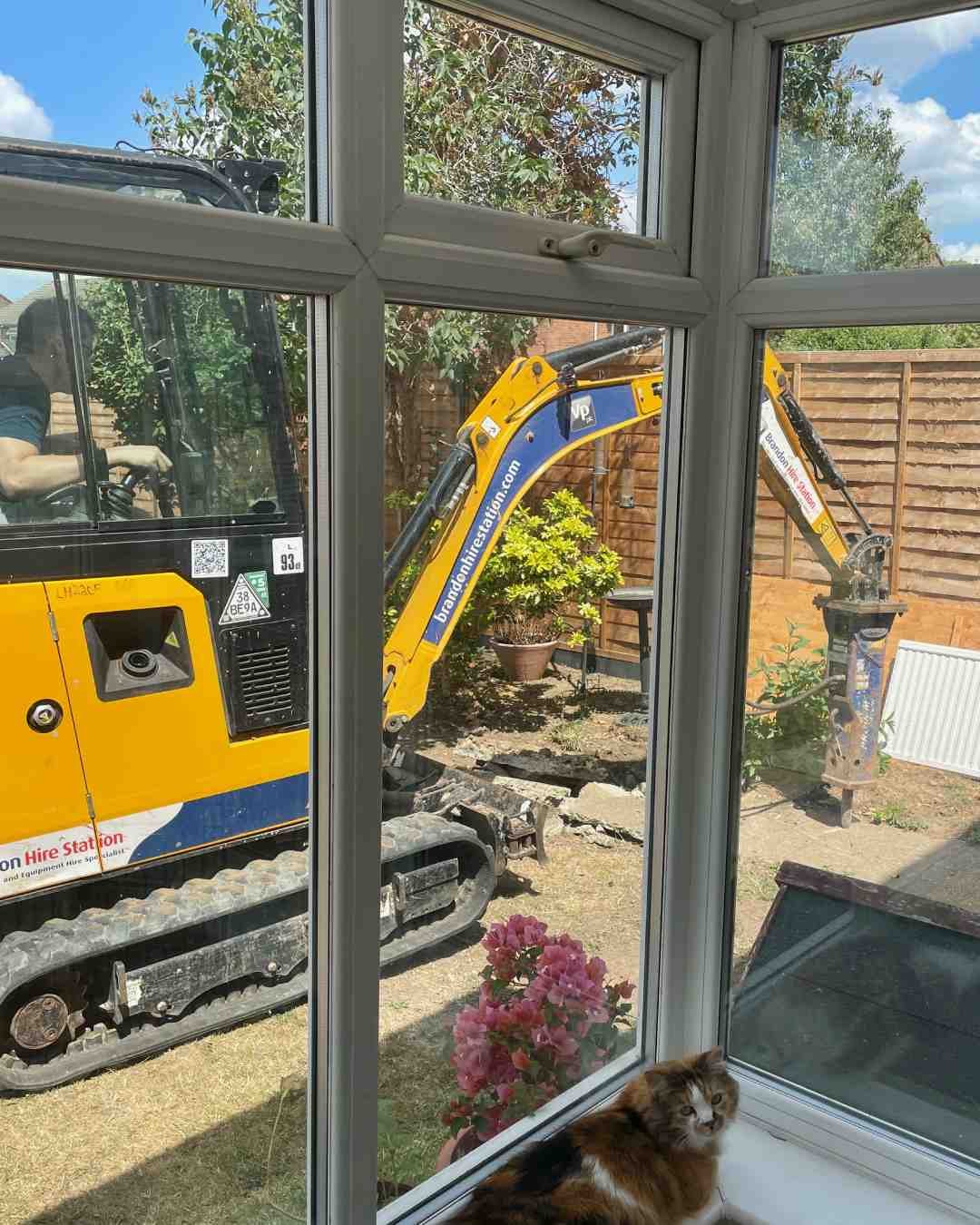 The Heavy Machinery Arrives Mini excavator with hydraulic breaker removing old concrete during our Surrey garden renovation, viewed from inside the conservatory.